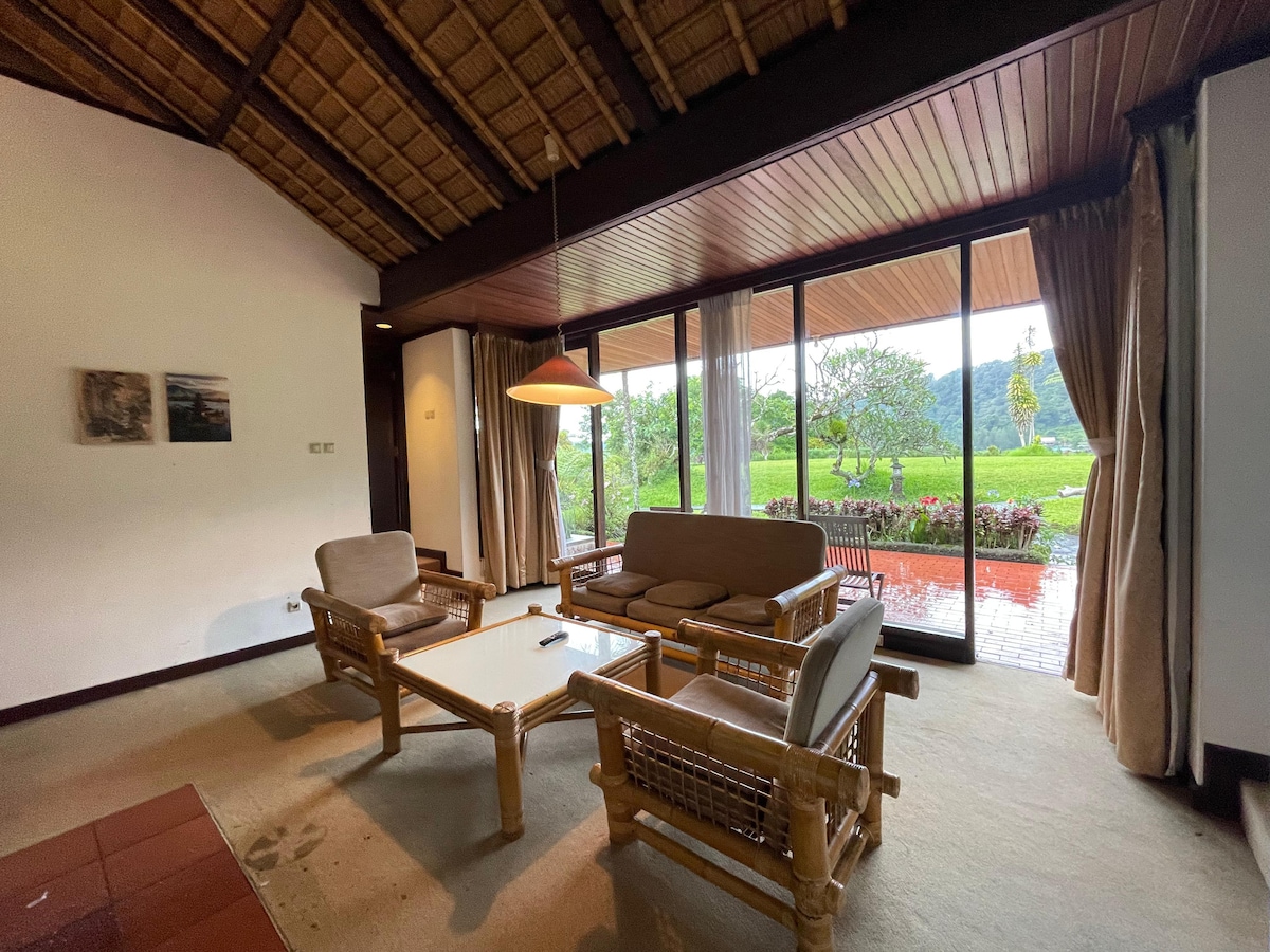 A cozy seating area is featured, showcasing a set of wooden furniture, including a rectangular coffee table and a spacious sofa. Large glass doors allow natural light to fill the room, while offering views of the green outdoors and a vibrant garden.