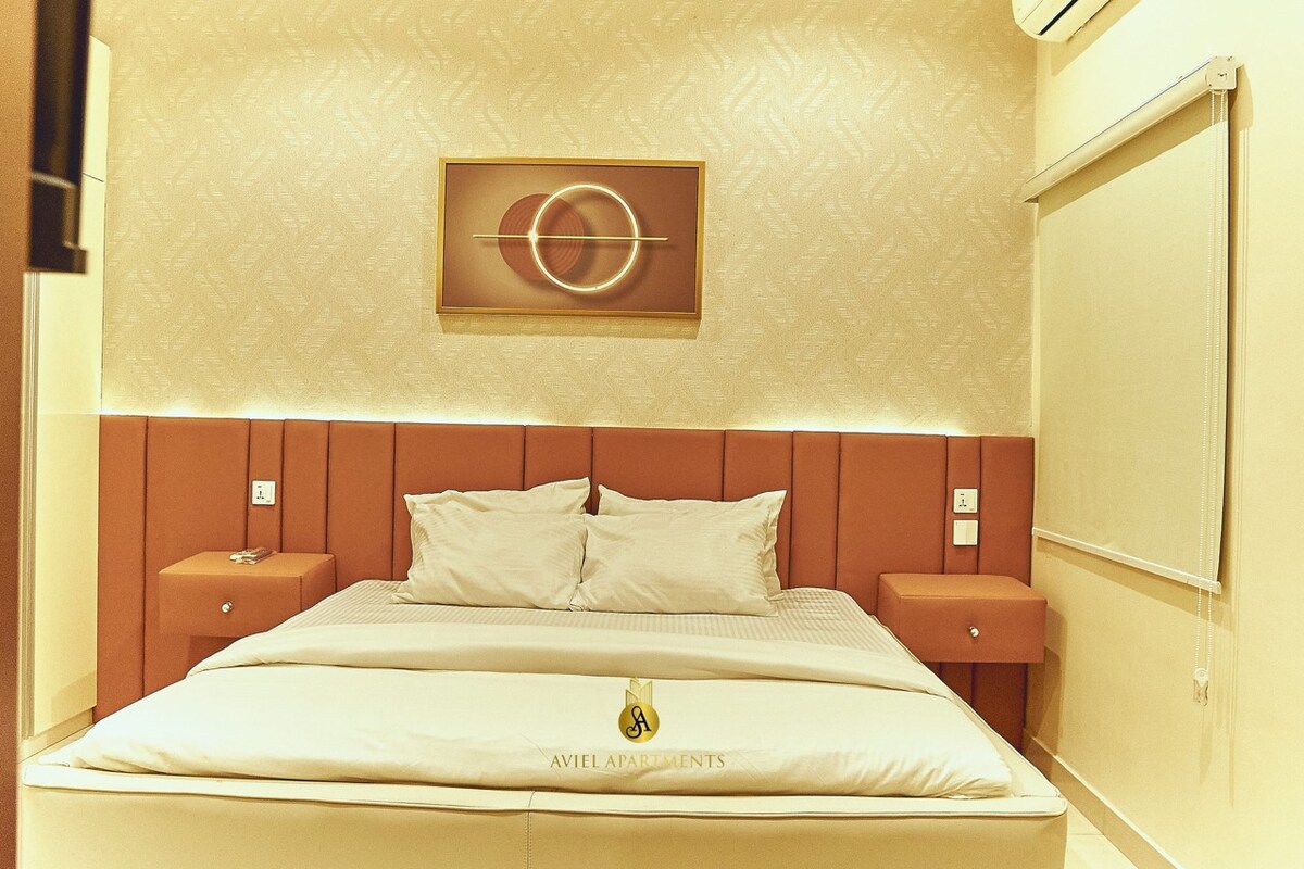 A serene bedroom features a spacious bed with plush white bedding and two bedside tables. A soft ambient light illuminates the room, highlighting the elegant wall decor. The design is complemented by a subtle patterned wall and a large window adorned with a light-filtering blind.