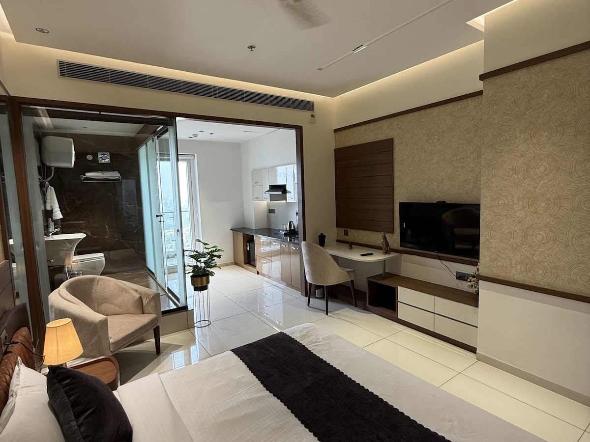A modern room features a spacious bed with neutral bedding, a cozy seating area with two chairs, and a television mounted on a smooth wall. The kitchenette is visible, along with large glass doors leading to a balcony. Natural light fills the room.