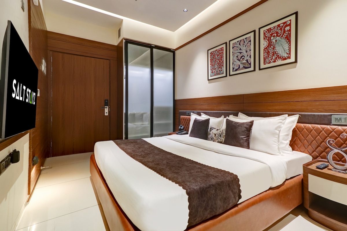A spacious room features a large bed with a brown and white bedding arrangement. The wall behind the bed is decorated with framed artwork. A television is mounted on the wall, and a glass partition leads to an adjoining lounge area.