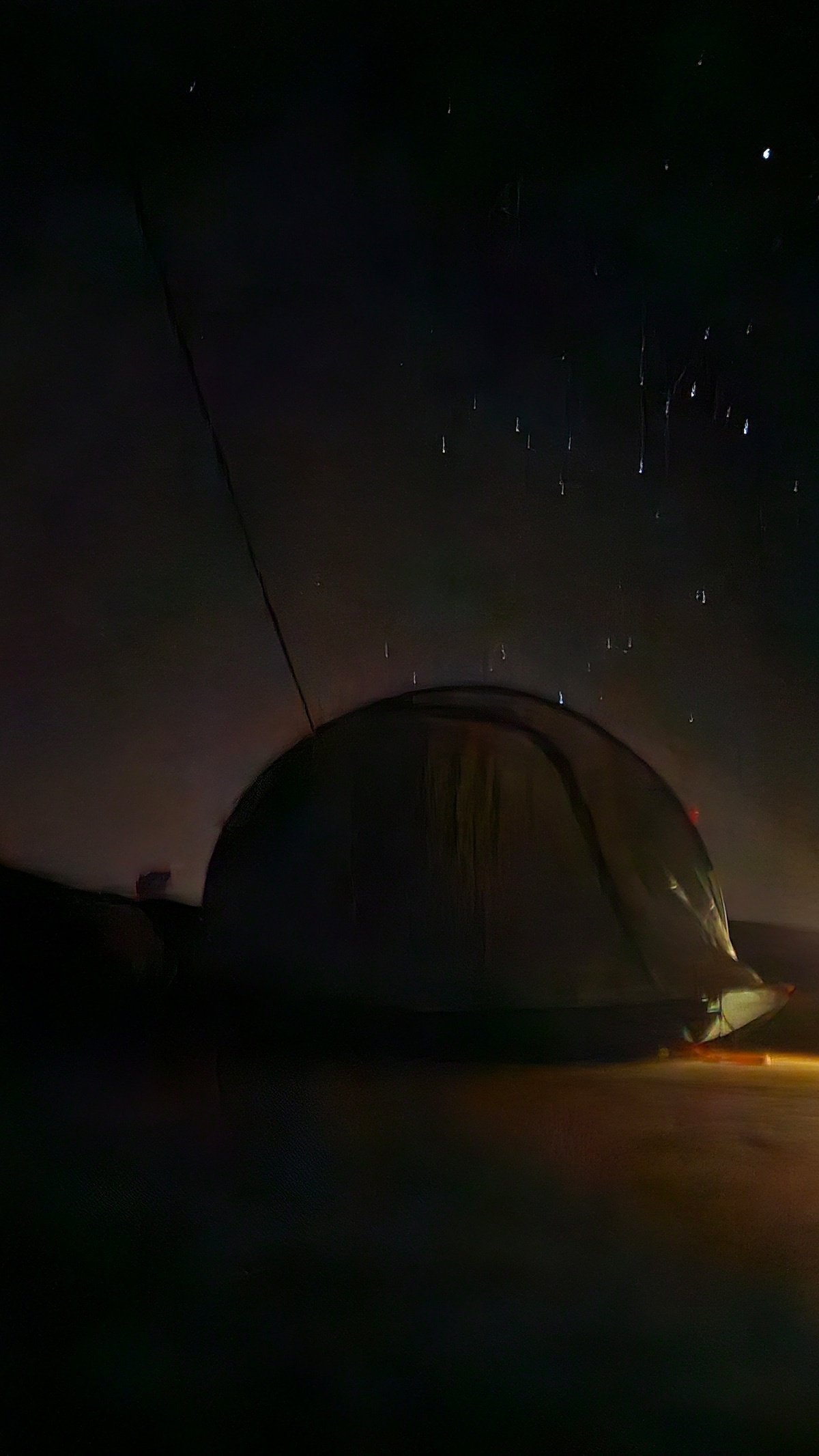 A camping tent is visible under a starry night sky, with faint streaks of light indicating falling stars. The scene is enveloped in darkness, highlighting the shape of the tent and a soft glow at its entrance.