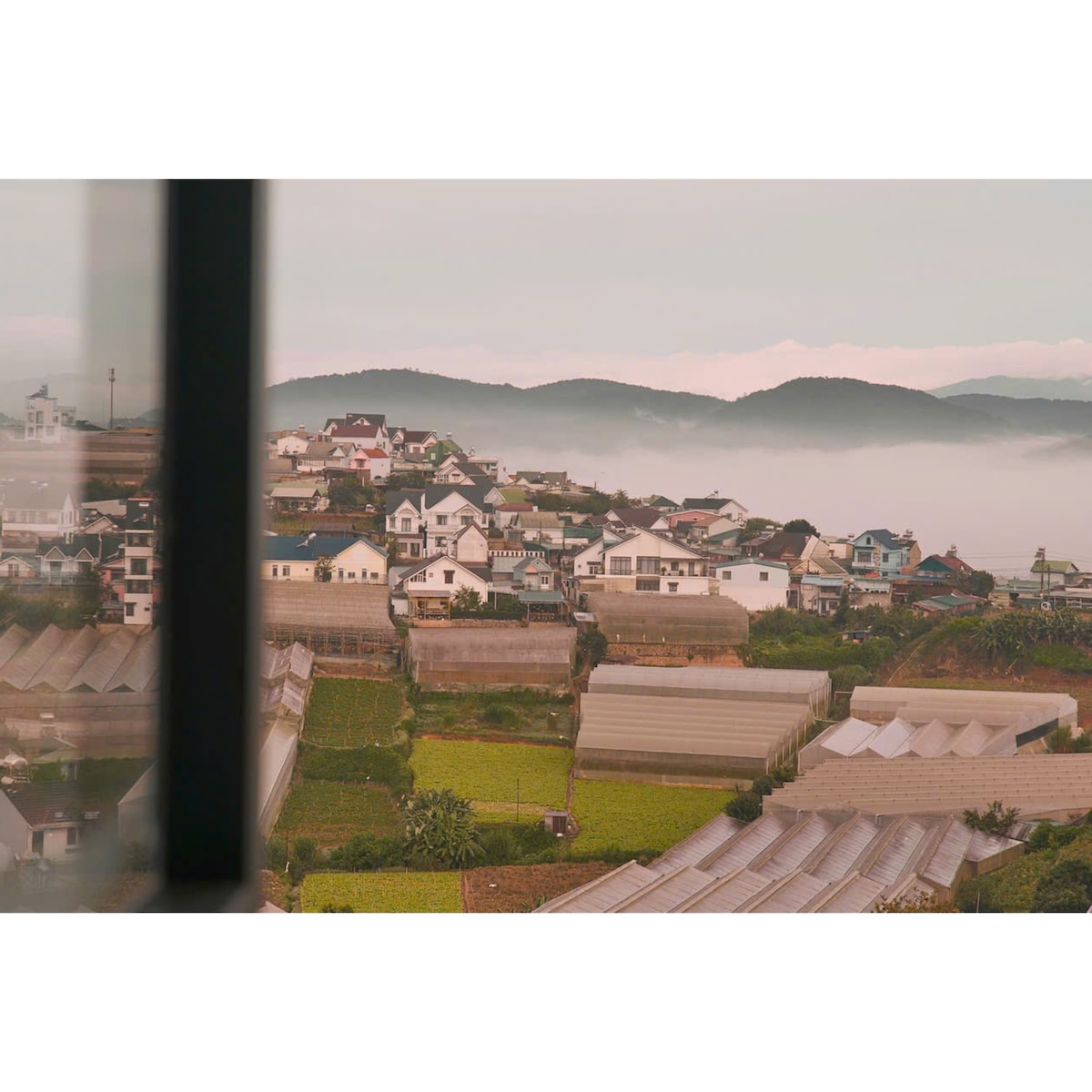 A view captures a serene landscape of houses nestled on rolling hills, shrouded in mist. Neighboring fields and greenhouses create a layered effect, while distant mountains rise softly in the background under a gentle light.