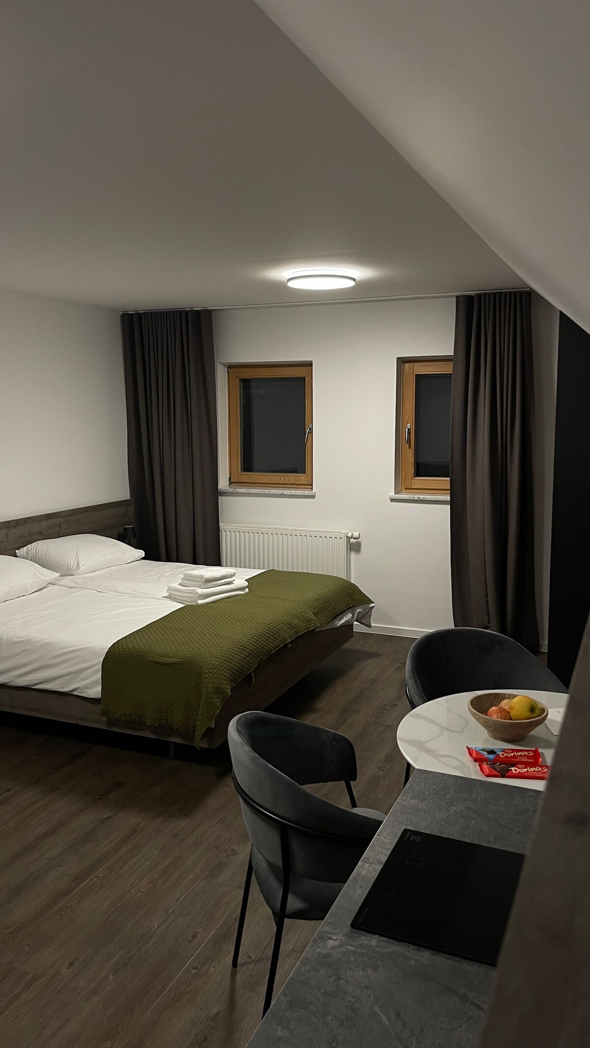 A well-arranged space features two single beds with white linens and a green throw at the foot. A small dining table is set with fruit. A kitchenette is visible with a sleek countertop. Two windows allow natural light to illuminate the room.