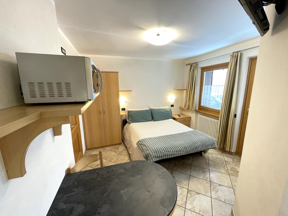The room features a spacious bed with light bedding, complemented by wooden cabinetry and soft lighting. A small microwave is positioned on a counter. A window adorned with light curtains allows natural light to enter, illuminating the tiled floor.