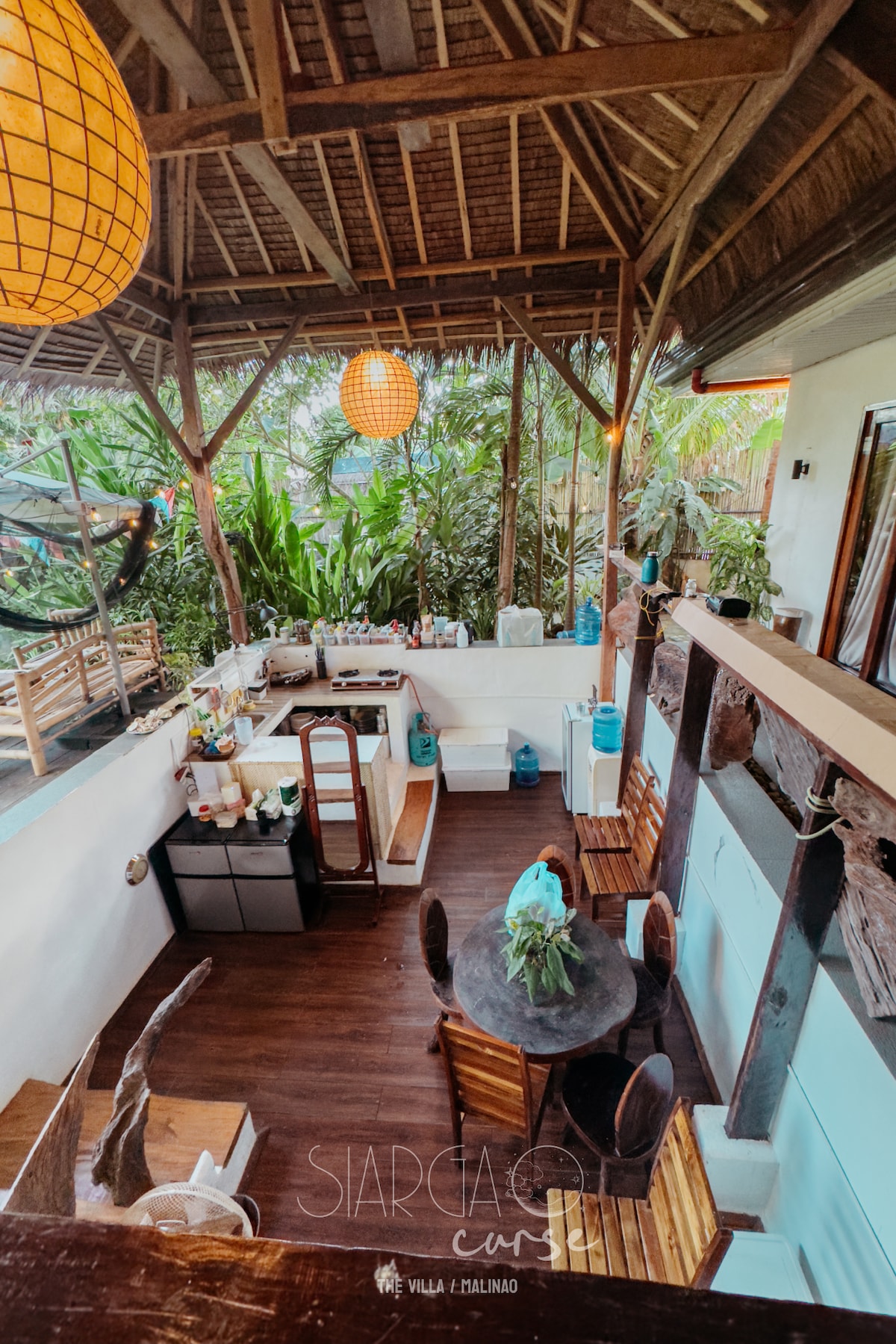 An open-plan kitchen and dining area features wooden furniture and tropical decor. A round table is set with chairs, and the kitchen space includes essential appliances. Large windows allow natural light to fill the room, while hanging lanterns add a warm glow amidst the surrounding greenery.