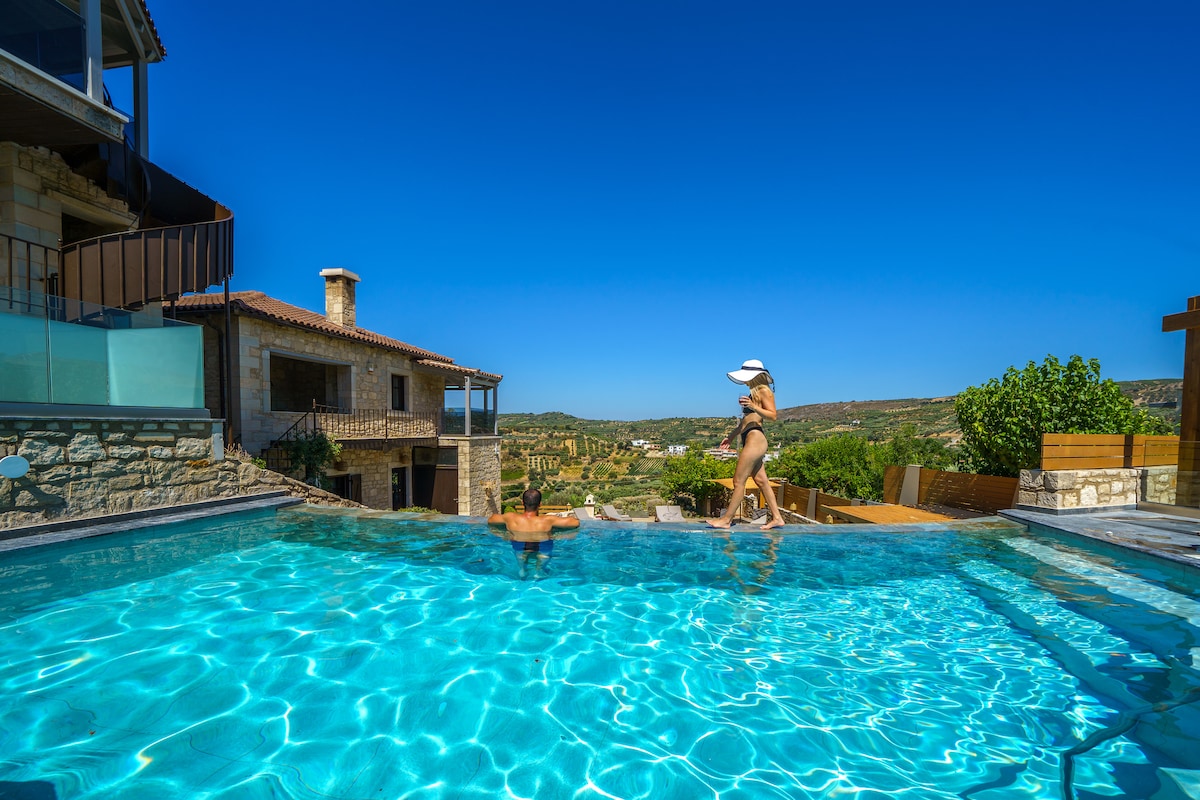 The private swimming pool glistens under a clear blue sky, surrounded by a sunlit stone terrace. The villa and scenic landscape are visible in the background, while lounging areas invite relaxation. The tranquil atmosphere emphasizes the inviting nature of this outdoor space.