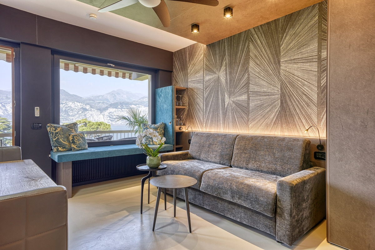 A modern living area is showcased with a comfortable gray sofa facing a large window that provides a scenic mountain view. Soft lighting creates a cozy atmosphere, and a round table is placed in front of the sofa. Decorative plants and cushions add a touch of elegance.