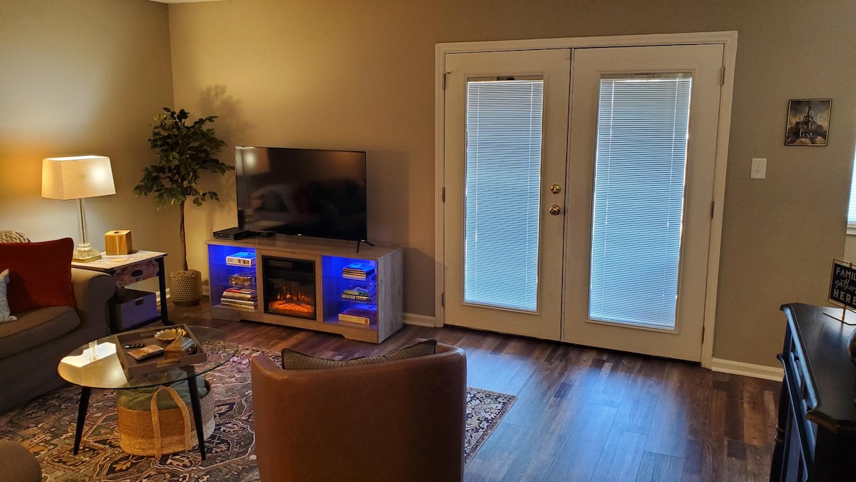 A cozy living area features a light brown sofa and an armchair, with a glass coffee table in front. A television is mounted above a modern entertainment unit, which has blue lighting. Two glass doors lead to an outdoor space, allowing natural light to enter.