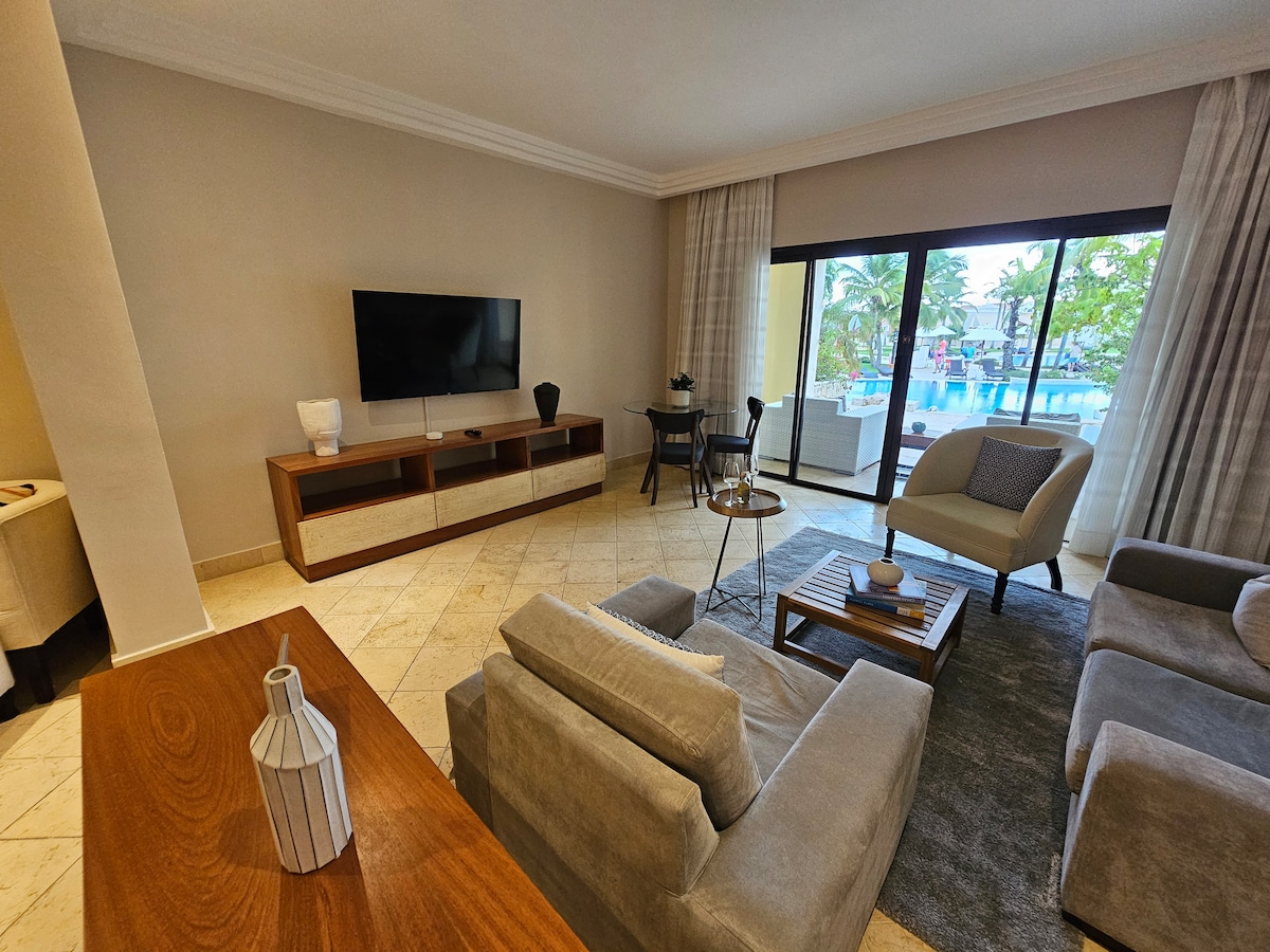 A spacious living area features modern furniture, including a light grey sofa and a sleek coffee table. A large television is mounted on a wooden media console. Natural light is filtered through sheer curtains, highlighting views of the resort-style pool and surrounding palm trees visible through expansive glass doors.