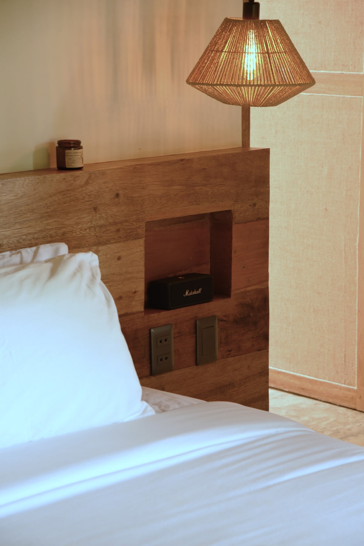 A close-up of a cozy sleeping area, featuring a wooden headboard with built-in storage. A stylish pendant light casts a warm glow, while a Marshall speaker sits within the recess. A spacious bed is adorned with white bedding, contributing to a serene atmosphere.