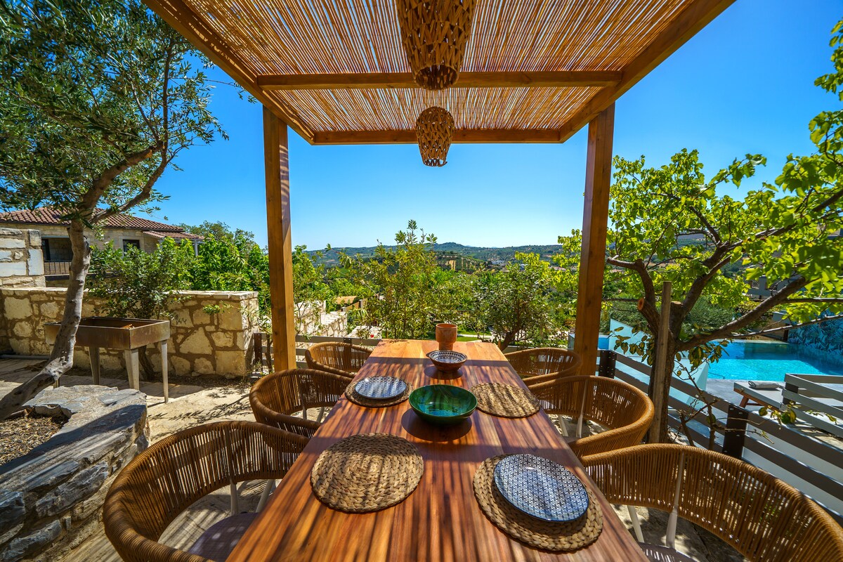 An outdoor dining area features a wooden table set with plates and bowls, surrounded by curved wicker chairs. A pergola above provides shade, with lush greenery visible in the background. Scenic views of the landscape and private pool enhance the relaxed atmosphere.