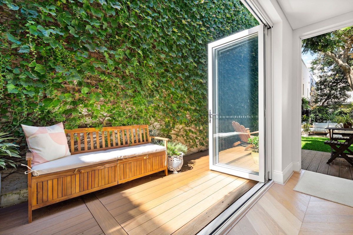A wooden bench is positioned against a lush green wall, providing a serene seating area. Large glass doors open to a sunny outdoor space, allowing for natural light to enhance the inviting ambiance.