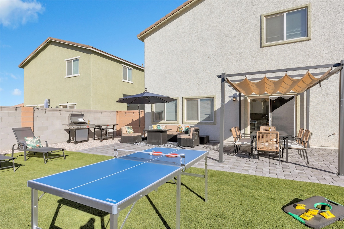 An inviting outdoor space is visible, featuring a Ping Pong table central to the grass lawn. Surrounding seating areas include a dining table and lounge chairs, all under a shaded pergola with earthy accents. The home’s neutral exterior complements the vibrant blue of the table.