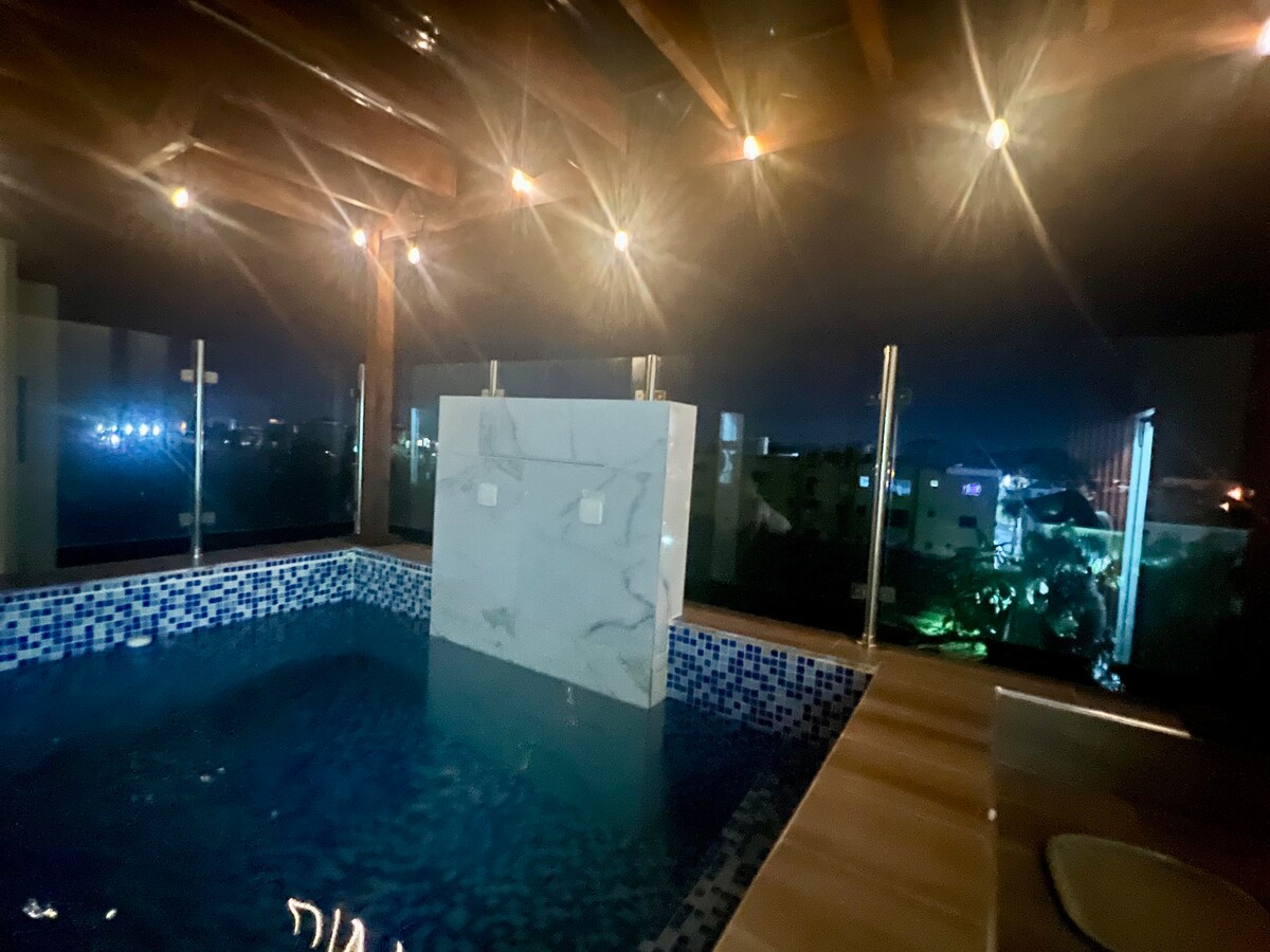 A private jacuzzi is showcased on a rooftop space, surrounded by a glass barrier for unobstructed views. The water reflects soft lighting, adding a serene ambiance. City lights can be seen in the background, contributing to a calm nighttime atmosphere.