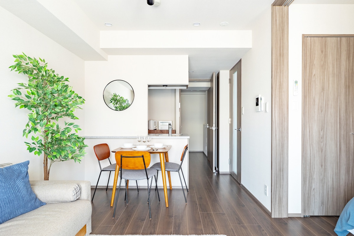 A modern dining area is featured with a round table and four wooden chairs, complemented by a decorative plant. The open layout allows for visibility into the kitchen area and a hallway leading to additional rooms.