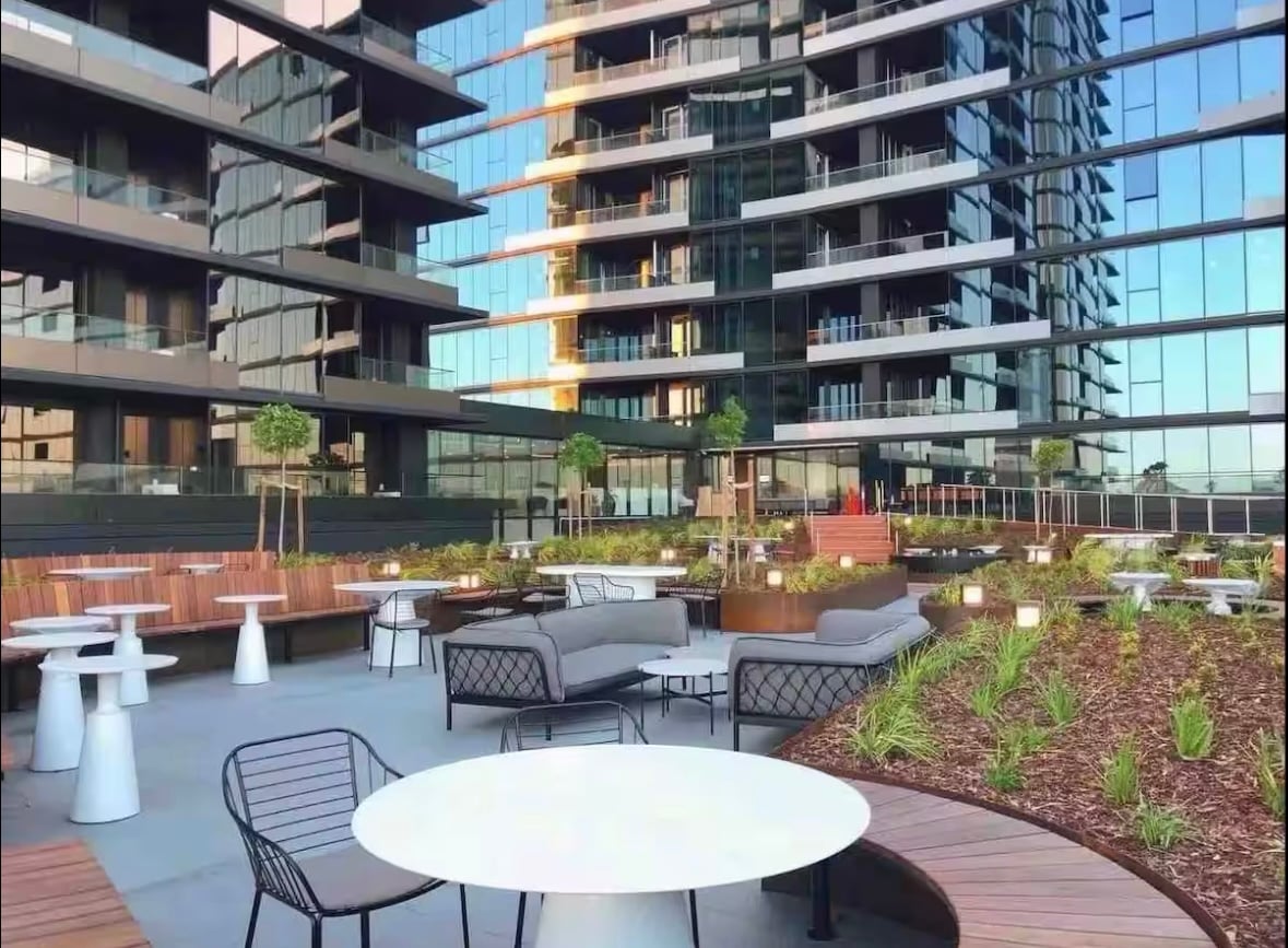 An outdoor communal area features a variety of seating options including circular tables and lounge chairs surrounded by sleek greenery. The modern building's glass facade reflects the sky, enhancing the space's ambience. Paved areas connect the seating to landscaped gardens.