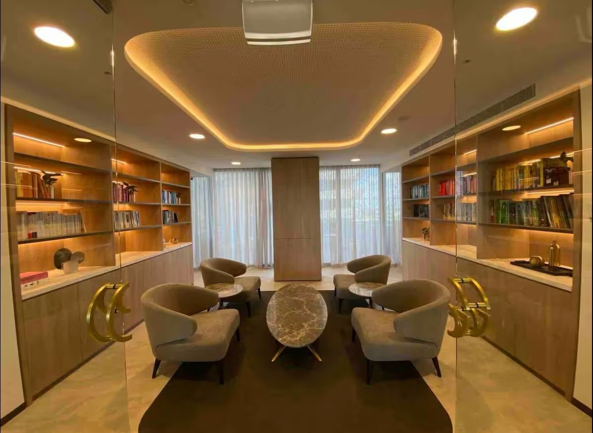 A well-lit communal lounge area is present, featuring a circular coffee table surrounded by four comfortable armchairs. Built-in shelves showcase a variety of books. Large windows allow natural light to fill the space, while a soft area rug adds to the room's warmth.