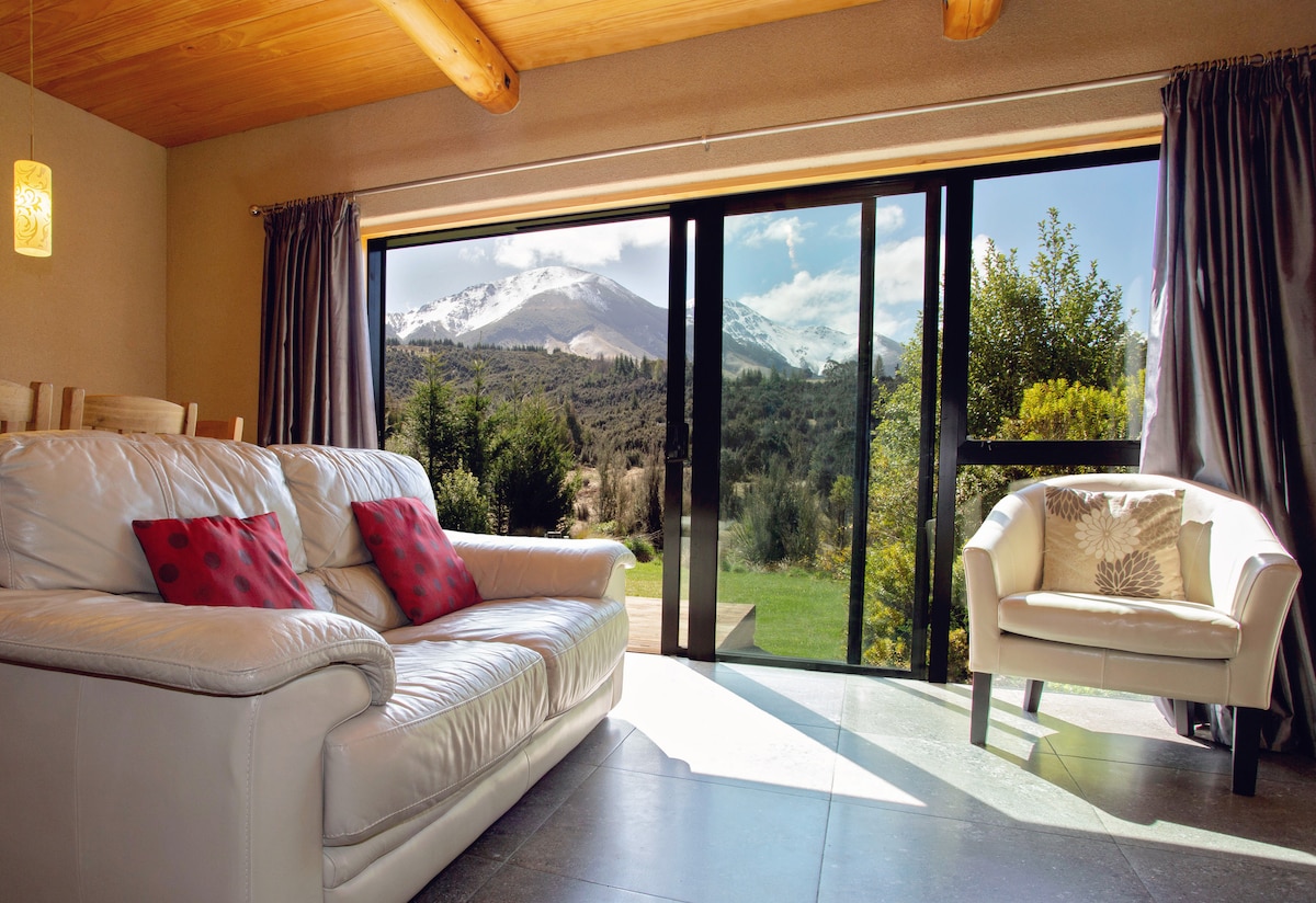 A comfortable lounge area features a leather suite with accent pillows, positioned to face expansive glass doors. Bright natural light floods the space, providing views of the surrounding mountains and lush greenery, enhancing the serene atmosphere.