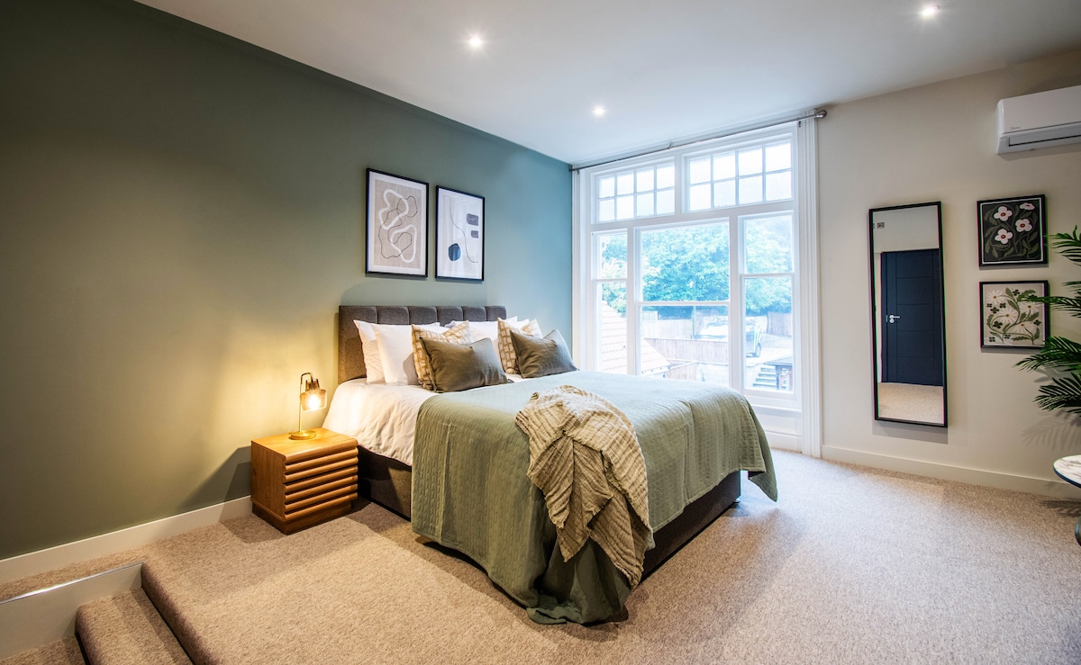 A spacious bedroom features a kingsize bed dressed in elegant linens and layered textiles. Large windows allow natural light to fill the room, illuminating a cozy armchair and a stylish mirror. Neutral tones on the walls enhance the serene ambiance of the space.