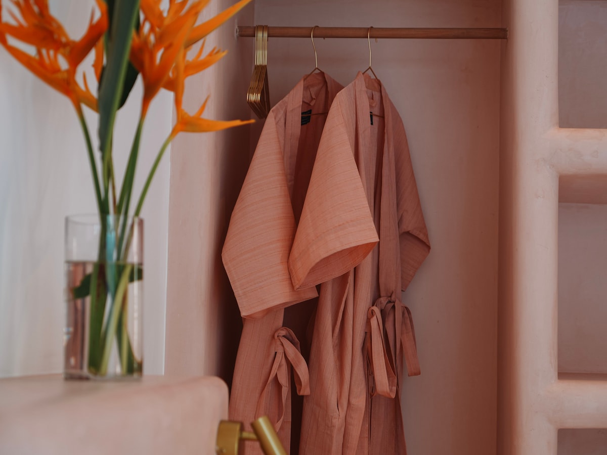Two soft, light pink robes hang neatly on wooden hangers inside a closet. A vibrant flower arrangement in a clear vase adds a touch of color, contrasting softly with the warm pink walls of the space.
