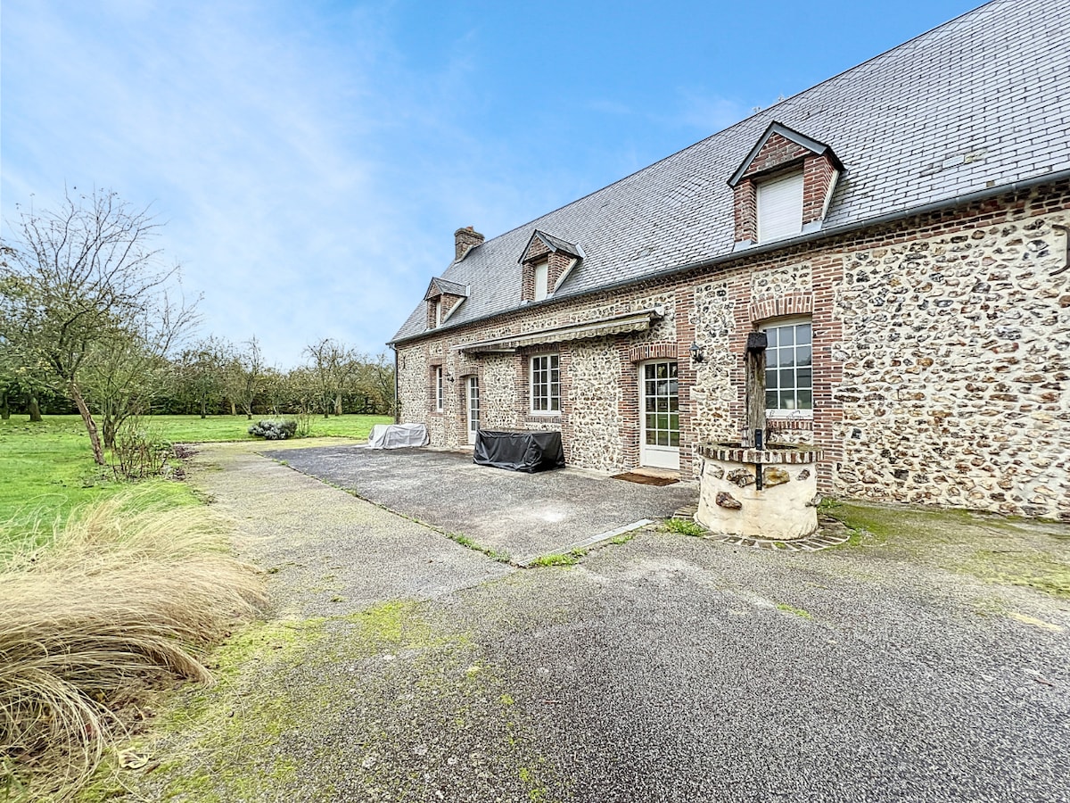 The exterior of a rustic stone house is depicted, featuring a spacious driveway leading to the entrance. Lush greenery surrounds the property, enhancing the natural setting. A covered area is visible, providing a sheltered space for outdoor activities.