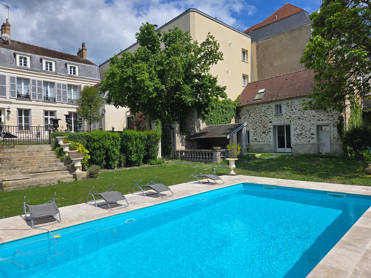 A tranquil outdoor setting features a heated pool surrounded by sun loungers. Lush greenery and trees provide shade, while the charming architecture of the nearby buildings showcases a blend of historical and modern elements. The scene is bright and inviting under a partly cloudy sky.