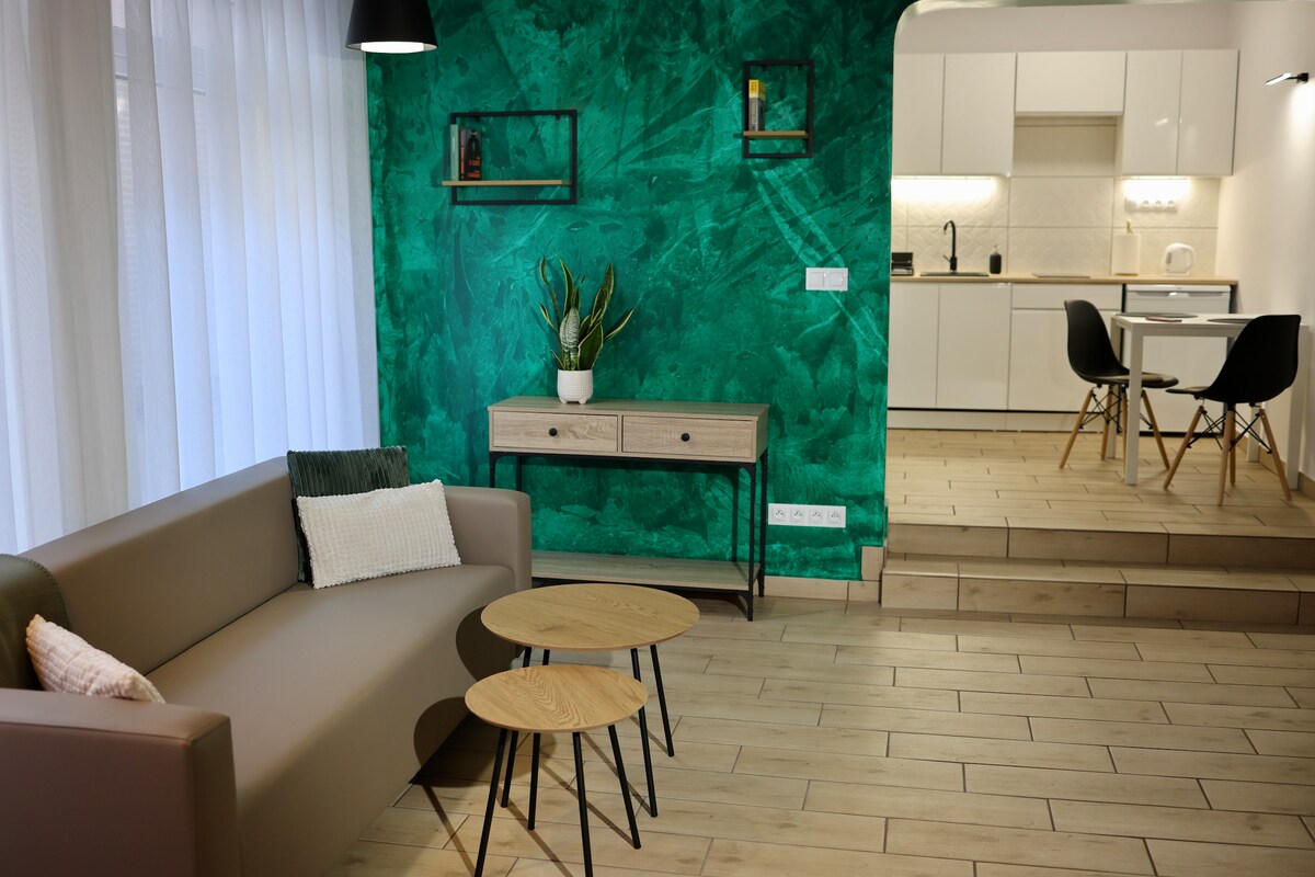 A modern living area features a green accent wall and a comfortable sofa. Two wooden side tables are positioned in front of the sofa. In the background, a kitchen area is visible, showcasing sleek cabinetry and a dining space with a table and chairs.
