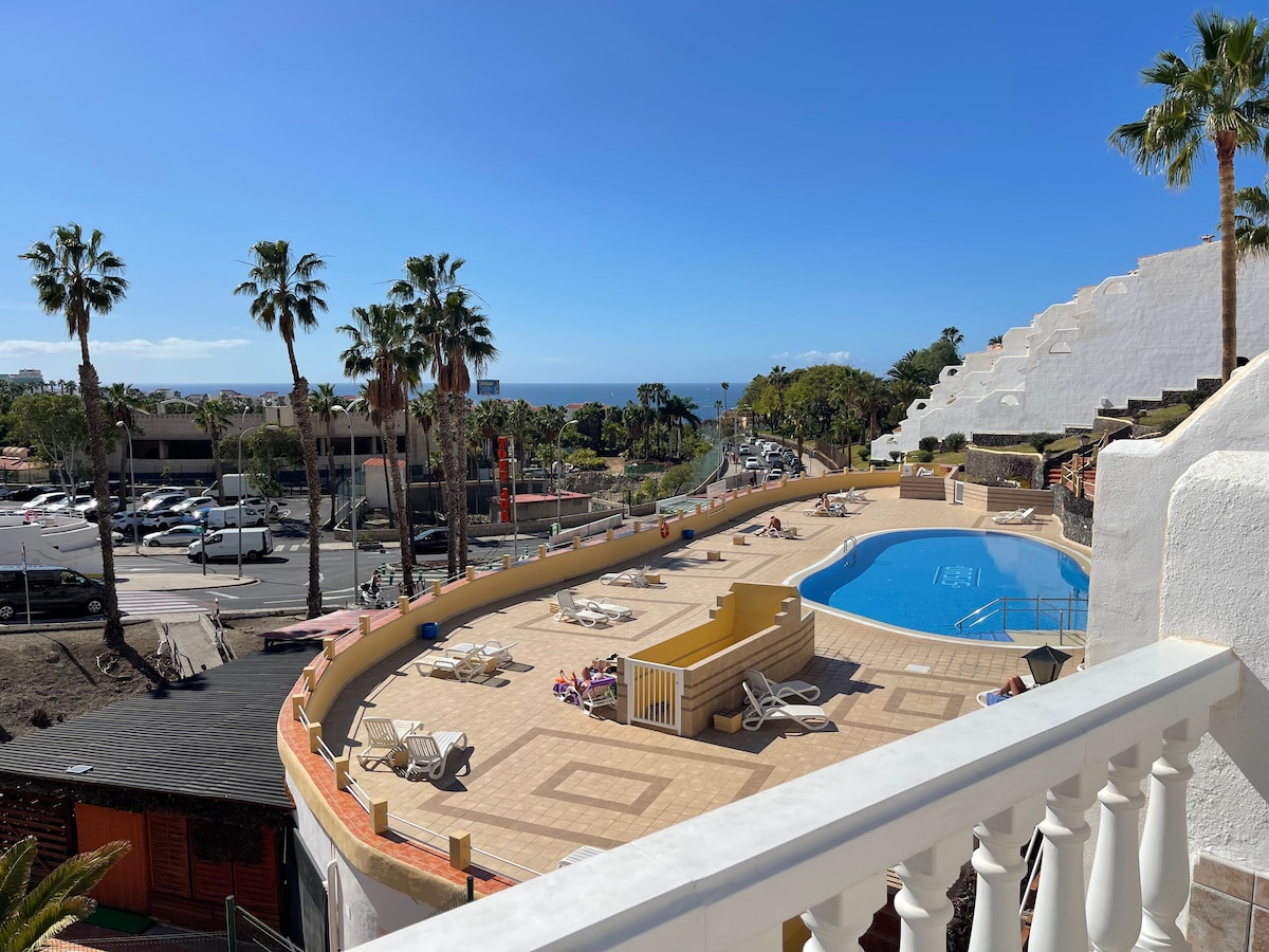 A view from the terrace features a communal pool surrounded by sunbeds, with palm trees standing nearby. The landscape includes a glimpse of the sea and the distant shoreline, providing a bright and open atmosphere. The area is well-maintained and inviting.