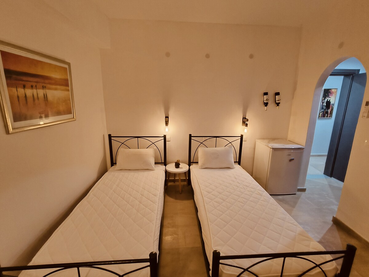 The image displays a bedroom with two slender beds arranged parallel to each other, featuring simple mattresses atop black metal frames. A small round table is placed beside one bed, while a white mini fridge is visible against a wall, enhancing practicality.