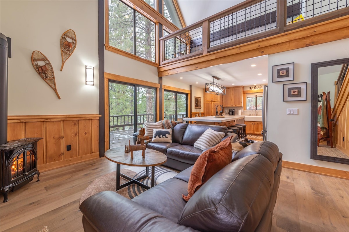 A spacious living area is showcased, featuring a dark leather sofa arranged around a circular coffee table. Natural light floods the room through large windows, highlighting wood paneling and inviting decor. A cozy fireplace stands to the left, contributing to the warm ambiance.
