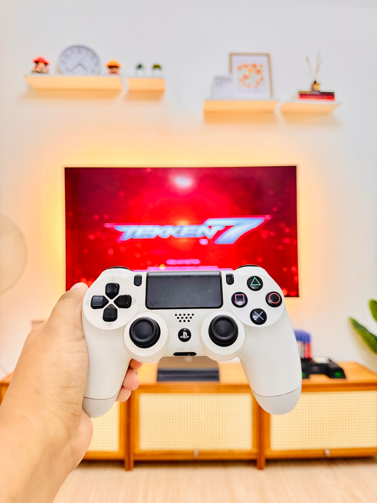 A gaming controller is held in front of a large television displaying a vibrant screen with a game title. A light wooden entertainment unit is visible below, complemented by decorative shelves and plants in the background, creating a lively gaming atmosphere.
