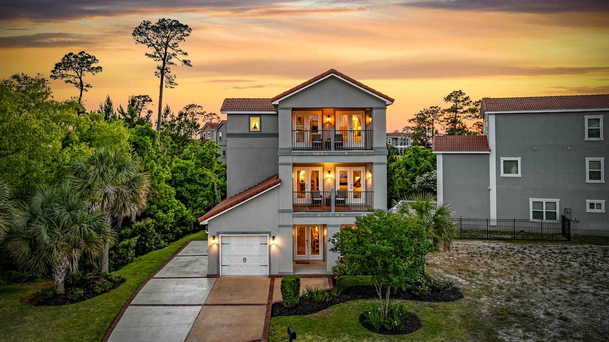 A modern three-story home is situated among lush greenery, visible under a vibrant sunset sky. Multiple balconies offer opportunities for outdoor enjoyment, while a driveway leads to a two-car garage. Palm trees and landscaped areas enhance the home’s welcoming appearance.