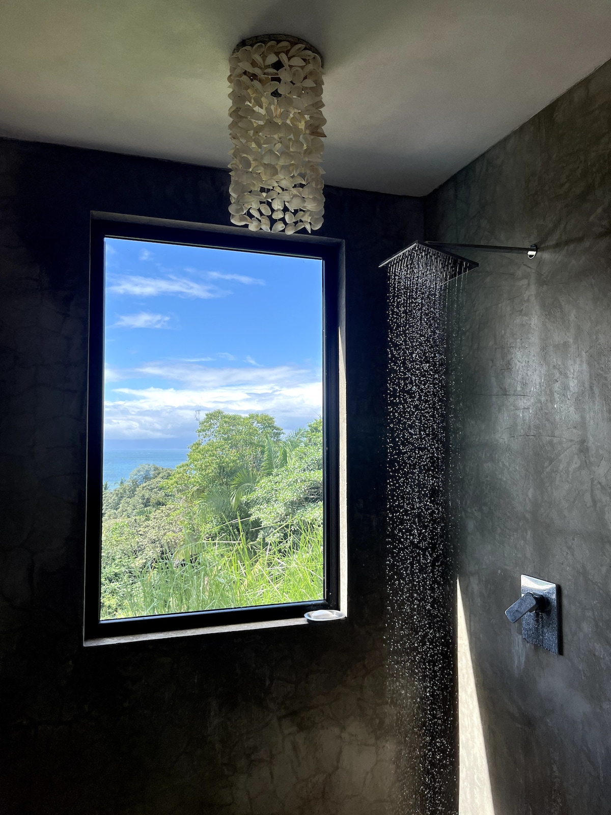 A modern shower area features a large window providing a view of lush greenery and a clear sky. Water flows gently from a sleek showerhead, while a unique light fixture adds a tasteful touch to the space.