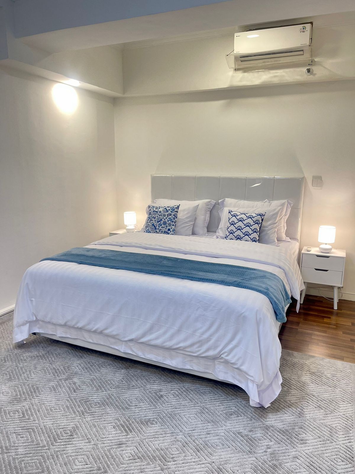 A bright bedroom features a spacious bed adorned with fresh linens and decorative pillows. Soft lighting from bedside lamps creates a cozy ambiance. Air conditioning is installed for comfort. A neutral carpet complements the wooden flooring.