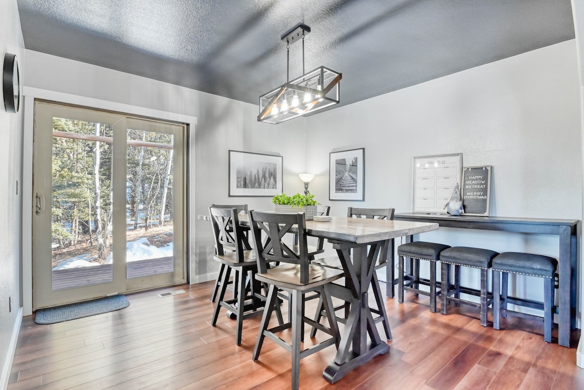 A dining area features a large wooden table surrounded by high-backed chairs, illuminated by a modern light fixture. Large sliding doors lead to a view of the surrounding woods. Wall art and decor add a stylish touch to the serene space.