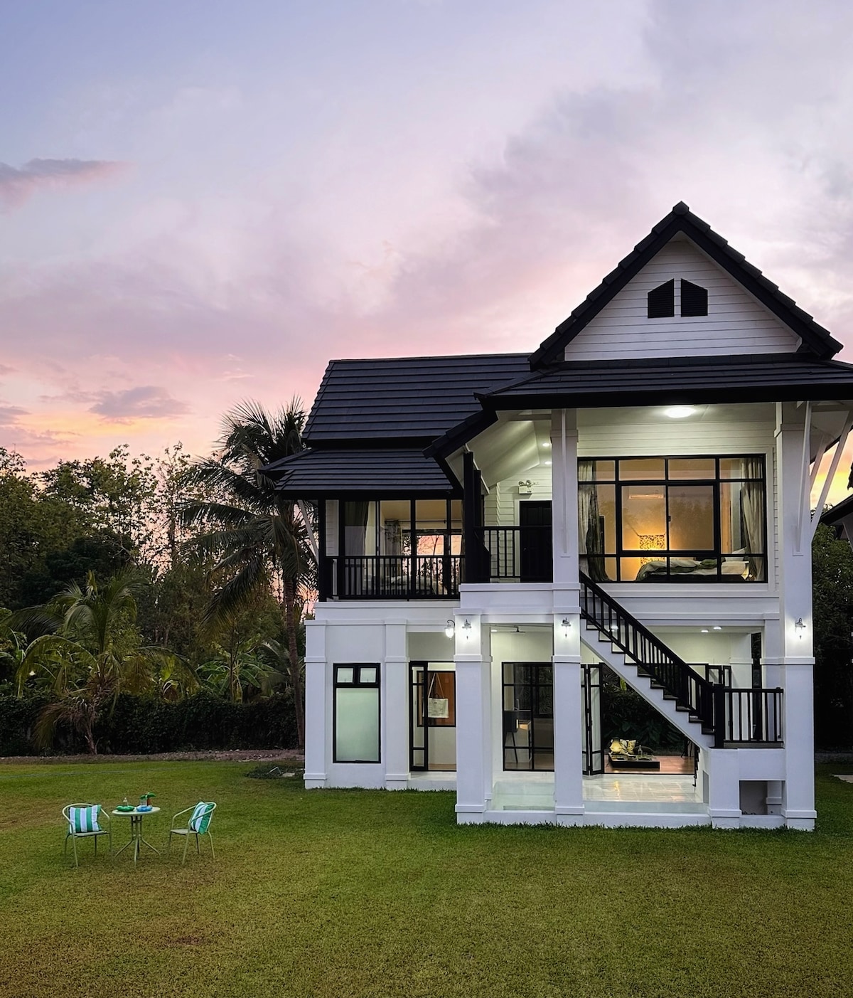 A two-story white house features large glass windows and a black roof. A staircase leads to the upper level. In the foreground, a small outdoor seating area with two striped chairs and a round table is situated on a green lawn, framed by trees.