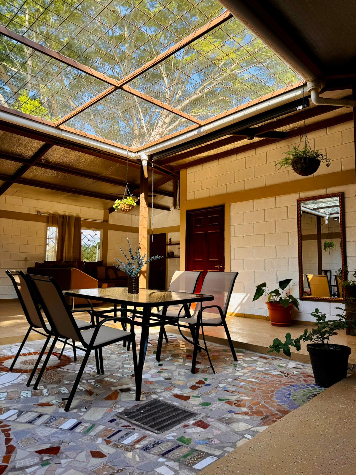An open interior space features a glass ceiling allowing natural light to fill the area. A mosaic floor is complemented by a simple dining table with chairs. Potted plants and wall mirrors enhance the sense of greenery and depth in this inviting setting.