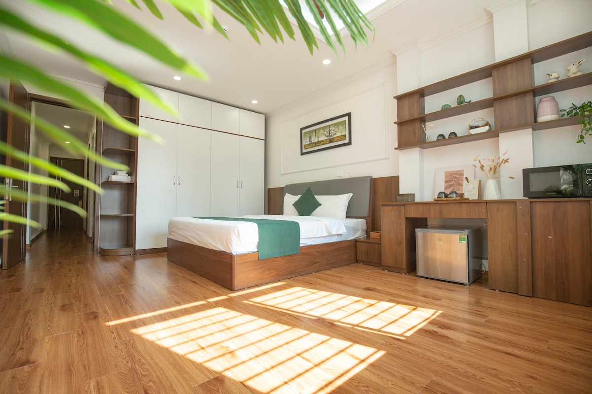 A spacious suite is presented with wooden flooring and neutral tones. Natural light fills the area, highlighting the queen-sized bed with a green throw, alongside a work desk and a mini-fridge. Built-in shelves display decorative accents, creating a calm, organized aesthetic.