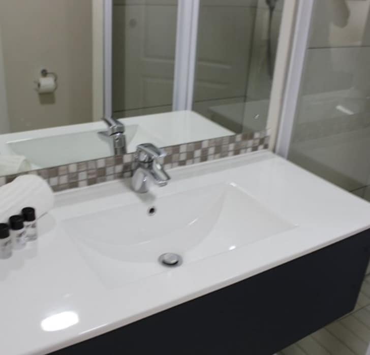 A modern bathroom features a sleek white sink with a minimalist faucet. A mosaic backsplash adds texture above the sink, while a large mirror reflects the space. Amenities include neatly arranged toiletries and a towel, enhancing functionality.