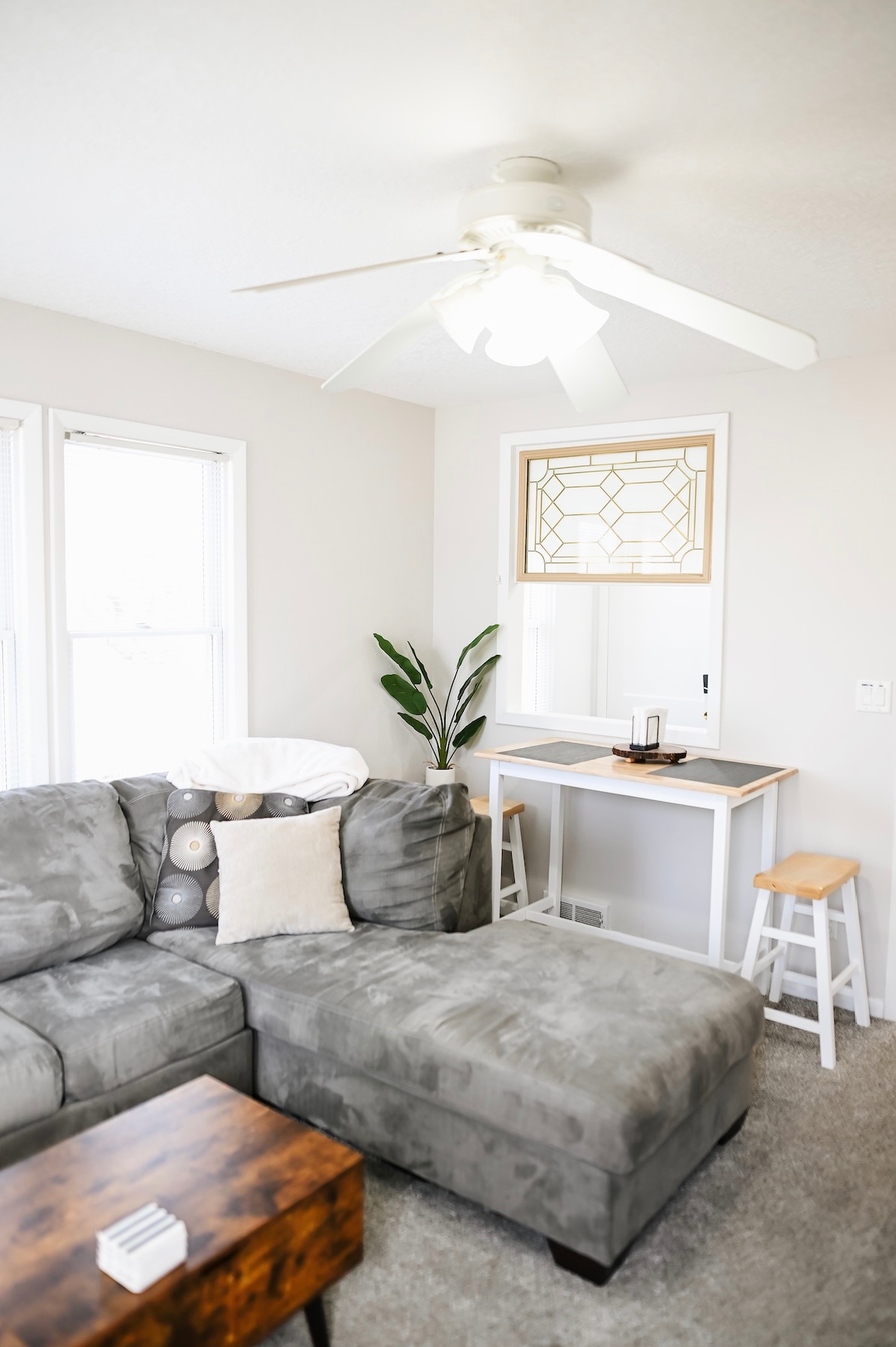 A comfortable living area features a plush, gray sectional sofa adorned with decorative pillows. A small, wooden coffee table rests in front. A light ceiling fan circulates air above, while natural light filters through nearby windows, highlighting a simple workspace with stools.