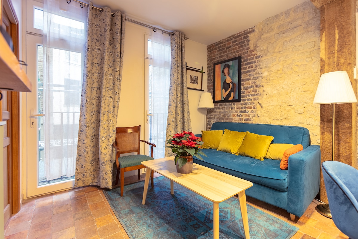 A cozy living room features a vibrant blue sofa adorned with yellow cushions, complemented by a wooden coffee table. A potted plant decorates the table, while light filters through the sheer curtains, highlighting the textured stone wall and framed artwork.