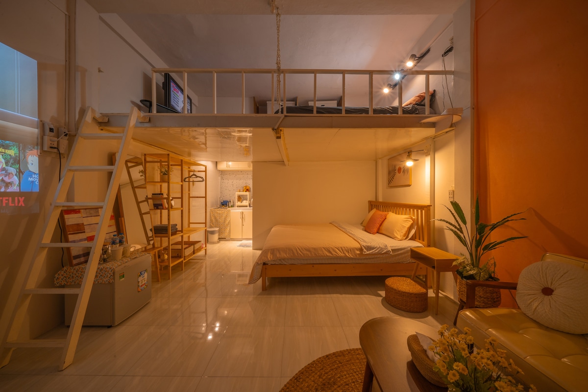 A cozy studio apartment is depicted, featuring a spacious bed and a lofted sleeping area. Natural light fills the room, highlighting a small kitchenette and seating area with a decorative plant. A projector screen is visible, indicating entertainment options, along with a contemporary, minimalist aesthetic.