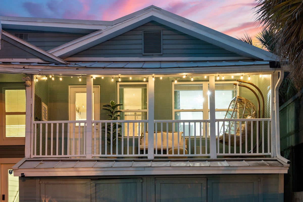 The exterior view of the guesthouse showcases a welcoming private balcony adorned with soft lighting. Two doors for entry can be seen, along with a suspended chair and a cozy seating area. The vibrant sky at dusk adds a gentle color contrast to the home.