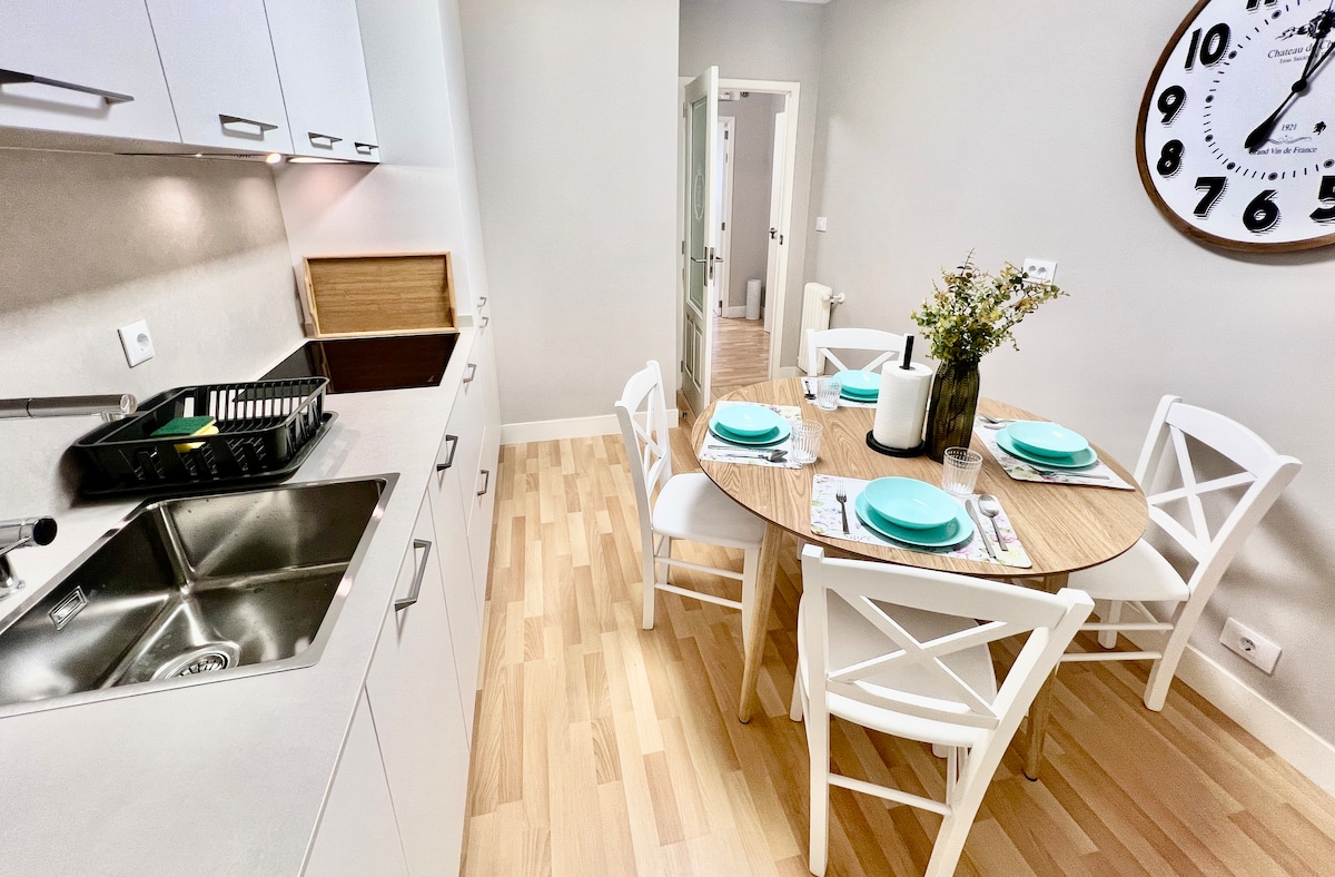A modern kitchen area features a stainless steel sink and a sleek countertop. A round dining table, set for four, displays turquoise plates and silverware. Light wood flooring complements the bright, neutral walls, contributing to a clean and functional design.
