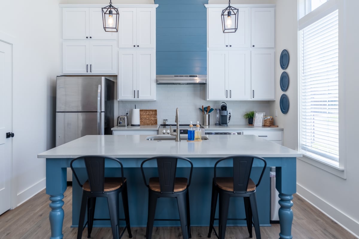 A modern kitchen showcases a spacious island with three black chairs. White cabinetry contrasts with a blue accent wall, and stainless steel appliances are visible. Natural light floods the area through large windows, brightening the inviting space.