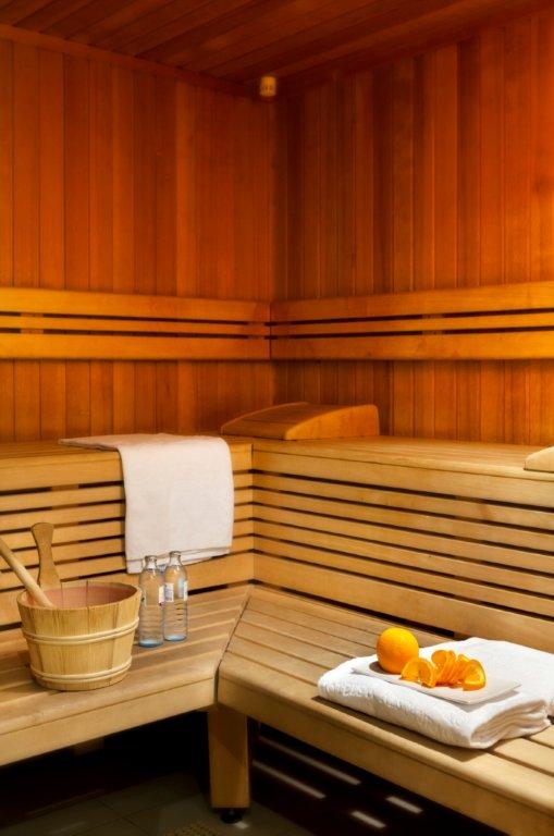 A wooden sauna features bench seating along the walls and is illuminated by warm lighting. A wooden bucket and ladle are positioned beside bottles of water, while fresh oranges are arranged on a towel. The natural wood grain creates a calming environment.