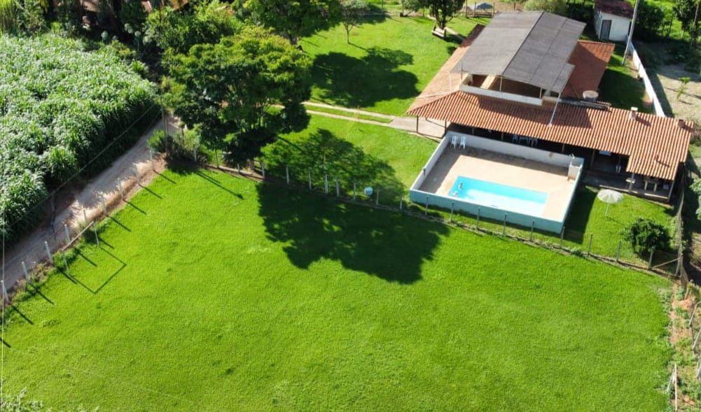 An aerial view reveals a spacious green yard surrounding a house with a terracotta roof. A rectangular swimming pool is set against the white wall of the outdoor area. Lush trees provide shade, while clear pathways lead to the house from the surrounding fields.