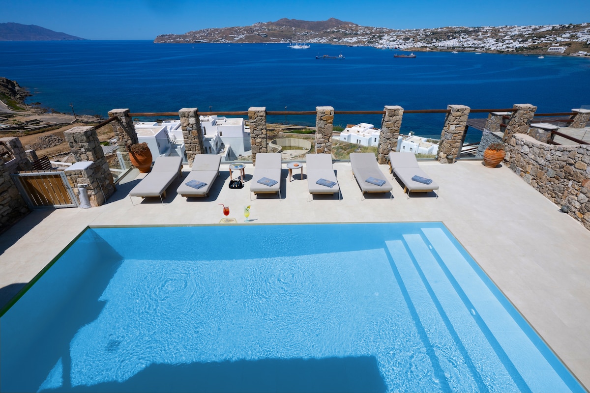 An infinity pool features a clear sky-blue surface, reflecting the surrounding scenery. Comfortable sun loungers are arranged along the poolside on a spacious terrace, offering unobstructed views of the sea and distant hills. The area is bordered by natural stone walls, enhancing the outdoor relaxation space.