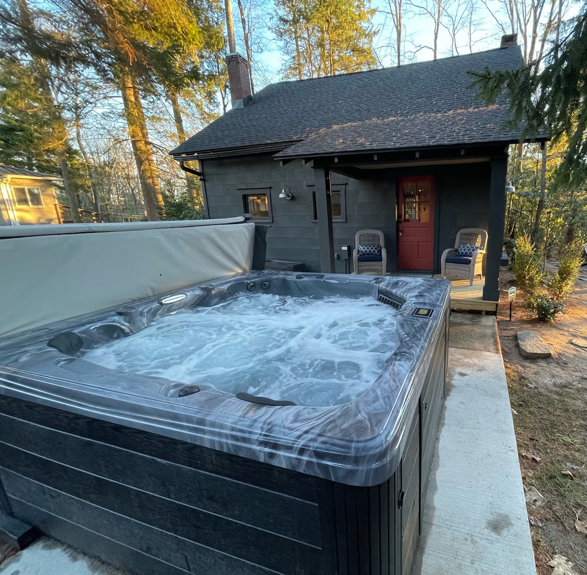 A spacious hot tub is situated outside the cabin, surrounded by a serene natural setting. The bubbling water invites relaxation, while the cabin's entrance, featuring a red door, is visible in the background. Two chairs are positioned nearby for comfortable seating.