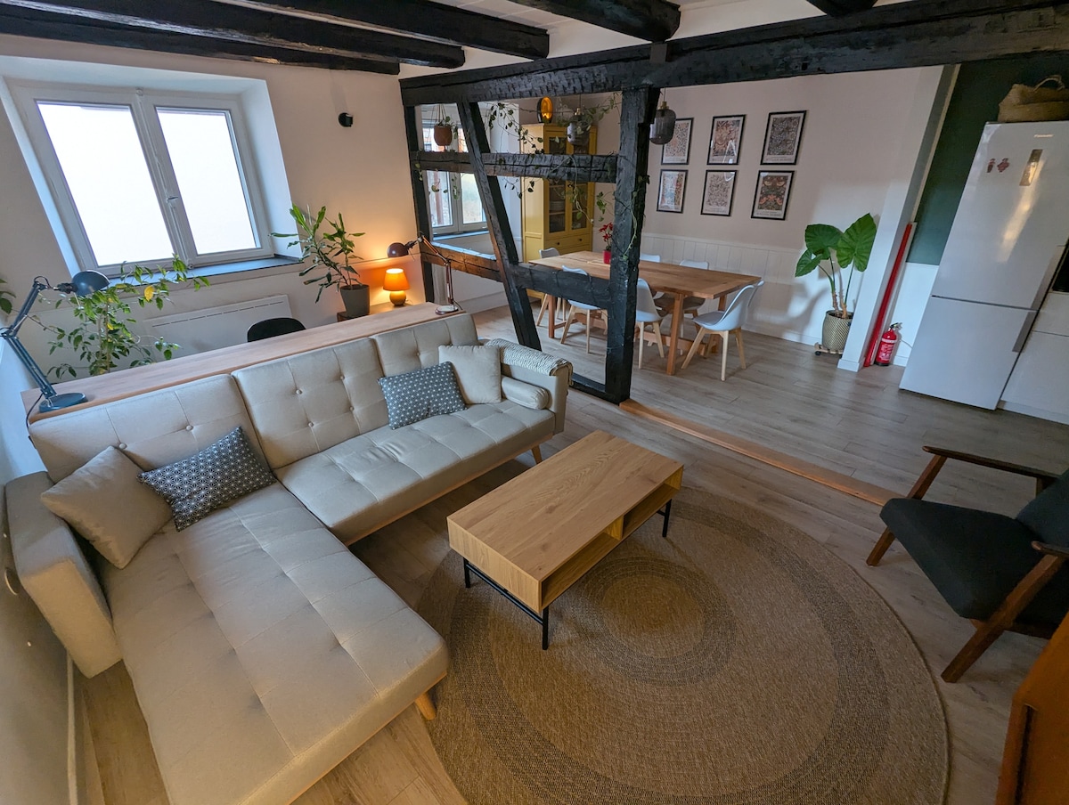 The living area features a light-colored sectional sofa and a wooden coffee table positioned on a round area rug. Two large windows allow natural light to enter the space. A dining area with a wooden table and chairs is visible in the background, along with houseplants and framed artwork on the walls.